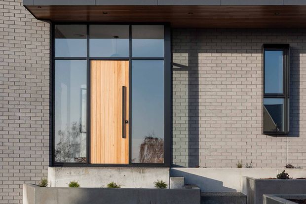 Modern residential front entry viewed from outside, featuring a tall natural timber pivot door framed by large dark metal glazing, pale brick masonry walls, a narrow vertical window, a projecting dark timber soffit, and minimalist concrete entry steps and planter walls.