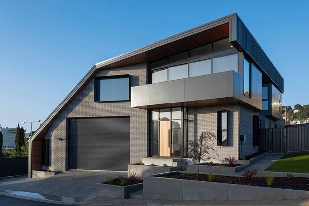 Contemporary two-storey residential house viewed from the street, featuring a steeply sloping side roof, dark grey brickwork, extensive glazing, a projecting upper-level balcony with metal cladding, and a large integrated garage. The front garden uses concrete retaining planters, dark mulch, and minimal ornamental planting.
