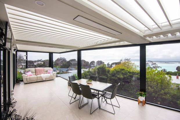 Bright, contemporary glazed living and dining area with expansive black-framed windows overlooking a coastal inlet or bay. The room features a tiled floor, a white sofa with pastel cushions, a dining table with dark chairs, and a striking slatted ceiling with integrated lighting and clerestory-style glazing.