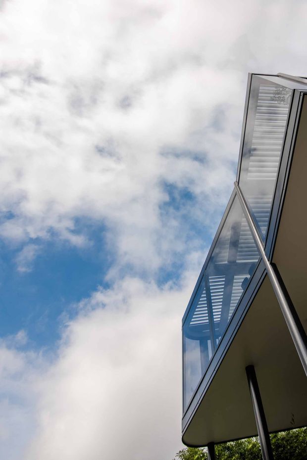 Low-angle exterior view looking up at a contemporary building with projecting balconies, transparent glass balustrades, slim metal supports, and horizontal louvres beneath the overhangs. The building edge contrasts against a partly cloudy blue sky, with a small amount of vegetation visible at the base.