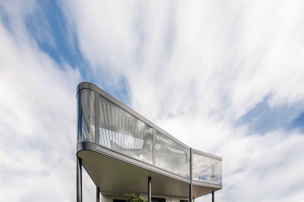 Low-angle exterior view of a contemporary multi-level building, focused on a projecting upper balcony or terrace with transparent glass balustrades, slim dark metal supports, and a curved metal-framed corner. The pale soffit and modern glazing stand against a bright, partly cloudy sky.