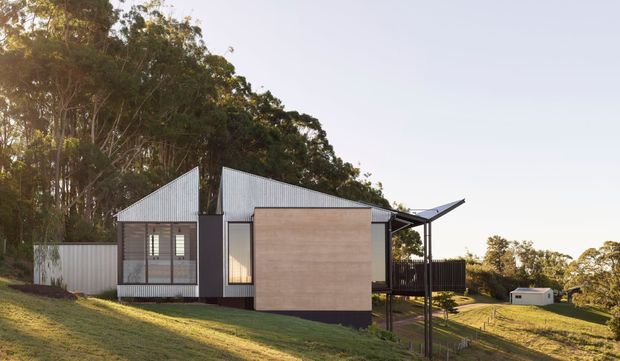 Exterior view of a contemporary single-storey house set on a sloping, heavily treed site. The home features angular pitched roof forms, corrugated metal cladding, dark vertical facade elements, timber-toned wall panels, large glazed openings, and an elevated deck or covered outdoor area.