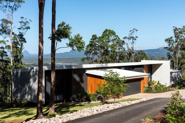 A contemporary single-storey house set into a wooded, elevated landscape. The building has crisp white rendered walls, dark vertical cladding, a low flat roof with a projecting angular canopy, and a warm timber-clad entrance or garage volume. A paved driveway and landscaped planting sit in the foreground, with expansive mountain and valley views beyond.