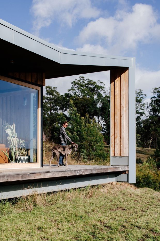 Exterior view of a compact contemporary building set in a grassy rural landscape. The composition highlights a projecting flat roof, open-sided covered deck or terrace, large glazed opening, warm vertical timber wall cladding, and pale grey metal exterior trim. Trees and a person with a dog are visible beyond the structure.