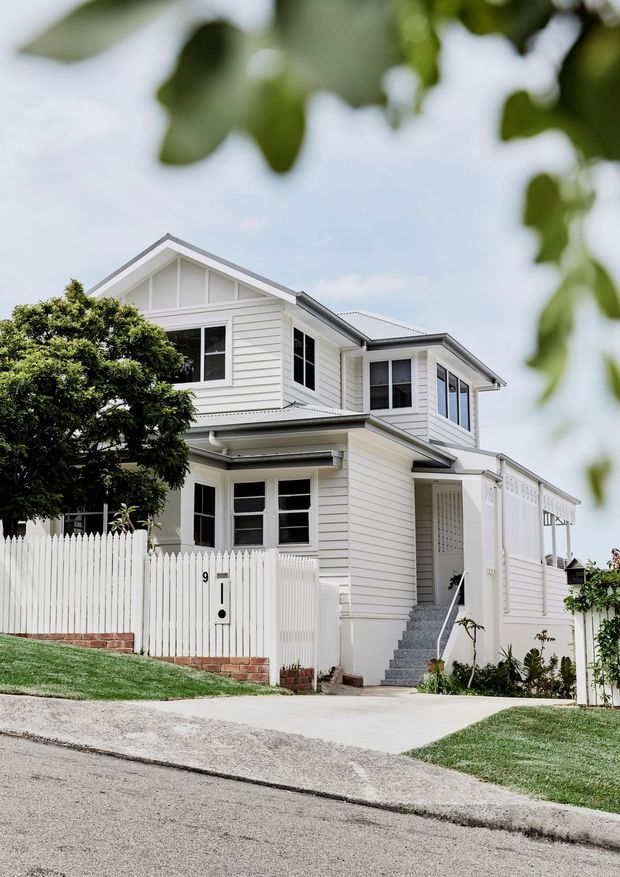 Exterior photograph of a two-storey white weatherboard home viewed from the street. The house features gabled roof forms, white-framed windows, a covered entry porch, front steps, a white picket fence, and a sloping suburban frontage.