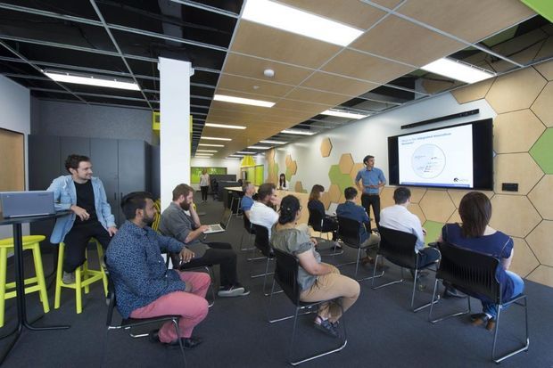 A contemporary commercial presentation or training room with attendees seated facing a presenter and projected screen. The space features an open-plan layout, dark carpet, exposed black ceiling services, suspended acoustic ceiling panels, linear lighting, white columns, and a geometric honeycomb-style timber-toned feature wall. Bright lime-green stools and accents add contrast.