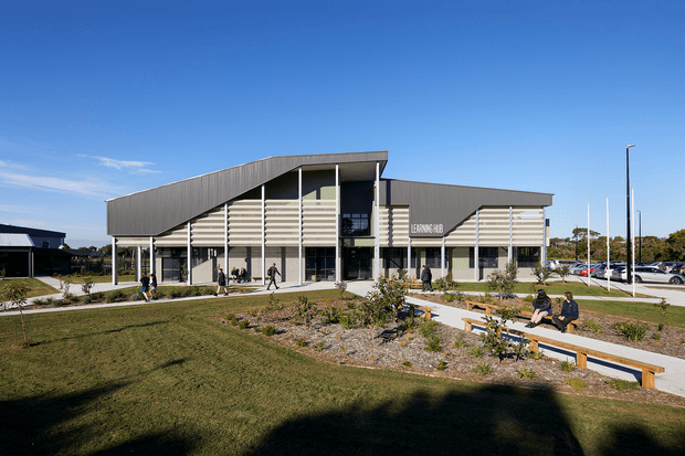 A contemporary multi-storey civic or commercial building viewed from a landscaped forecourt. The building has a dramatic sloping roof form, extensive horizontal louvres, exposed structural columns, large glazed entrances, and a visible 'Learning Hub' sign. Paved walkways, lawns, garden beds, benches, pedestrians, and a parking area surround the building under a clear blue sky.