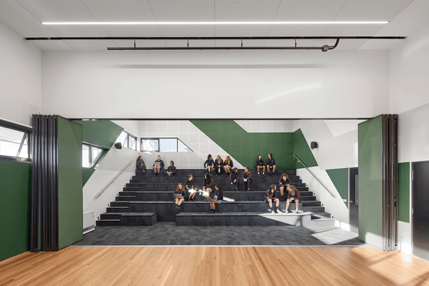 A bright contemporary multi-purpose communal interior with tiered black upholstered bench seating arranged like an informal auditorium or lecture space. Students are seated across the stepped platforms, with green and white geometric wall panels, exposed black services, acoustic ceiling panels, large windows, and a broad light timber floor in the foreground. Retractable dark partition walls frame the space.