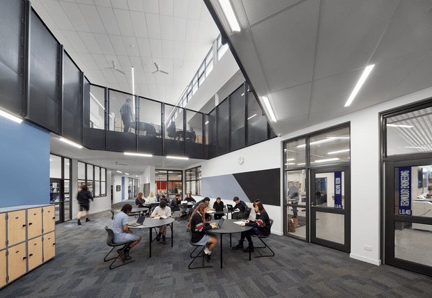 A bright, contemporary two-level communal interior resembling a school or institutional student commons. The open ground-floor area contains multiple round tables with people seated, surrounded by dark acoustic or glazed upper-level balustrades and visible rooms beyond. Large internal windows and glazed doors, linear LED lighting, ceiling fans, grey carpet tiles, white walls, and blue geometric wall accents create a clean modern educational environment. Fixed light-wood locker cabinetry is visible along the left wall.