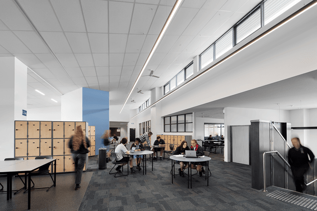 A bright, contemporary multi-level commercial interior resembling a school, university, or shared workplace. The open-plan space features long communal tables, round café-style tables, people working, extensive timber locker storage, a blue feature wall, dark patterned carpet, white walls and ceilings, high clerestory windows, linear LED lighting, and a stairwell with metal balustrades. The double-height volume and upper-level glazing create an airy, collaborative environment.