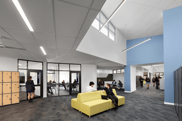 A bright, spacious contemporary commercial interior, likely a school or institutional common area, with a large open-plan lounge and circulation space. The room features a yellow sectional sofa, grey patterned carpet, blue and white walls, black-framed glazed meeting or classroom rooms, timber lockers, café-style tables, and multiple groups of people. A high ceiling with clerestory glazing, exposed white structural surfaces, suspended acoustic ceiling panels, ceiling fans, and linear LED lighting creates an airy, functional environment.