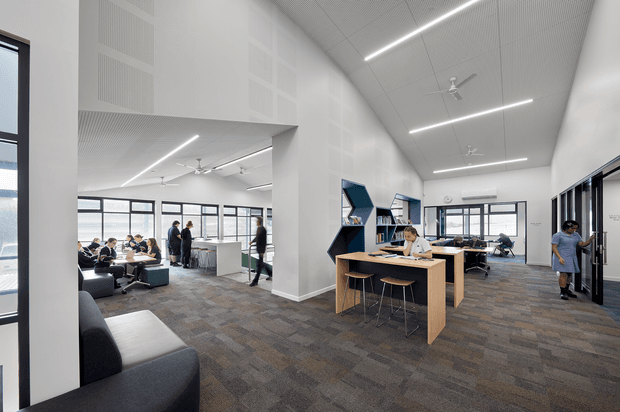 Bright, open-plan contemporary workspace with desks, shelving, a central timber work counter, lounge seating, and multiple collaborative seating areas. The room has high ceilings, large black-framed windows, exposed acoustic ceiling panels, linear LED lighting, ceiling fans, and grey patterned carpet. People are working and walking through the space.