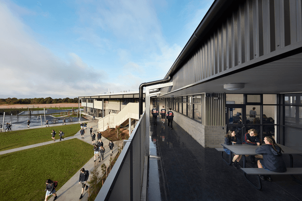 Exterior elevated view of a contemporary multi-storey institutional building, showing a long covered balcony or circulation terrace with dark metal framing, extensive glazing, vertical facade fins, and light brickwork. People gather at outdoor picnic tables, while landscaped grounds, walkways, and a waterfront or sports-field-like area are visible beyond.