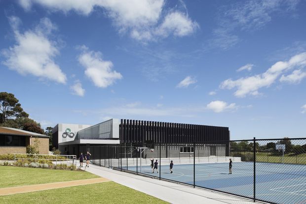 Exterior view of a contemporary sports facility beside a fenced blue tennis court. The building has a dark vertical slatted facade, light rendered or metal-clad volumes, extensive glazing, and a gently angled roof form. Landscaped lawns and paved pedestrian paths lead toward the entrance, with a bright blue sky and neighboring low-rise structures in the background.