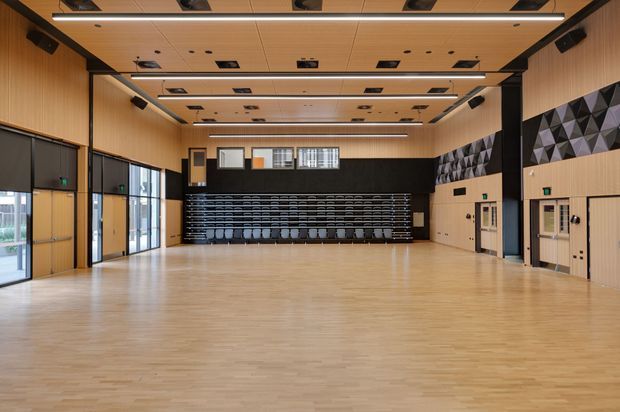 A large, bright multi-purpose hall or auditorium with a polished light timber floor, timber acoustic wall and ceiling panelling, black structural framing, high clerestory windows, retractable or stacked seating at the rear, and geometric acoustic panels on the right wall. The space appears designed for assemblies, performances, presentations, or community events.