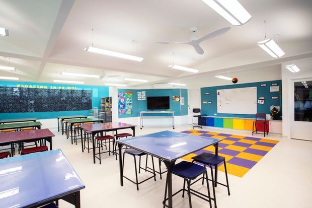 Bright, spacious modern classroom with grouped student tables and chairs, turquoise feature walls, a large whiteboard, interactive display, colourful built-in storage, and a bold purple-and-yellow patterned floor area. Linear fluorescent lighting and ceiling fans create a functional educational environment.