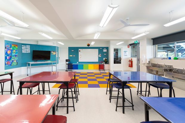 Bright, spacious multi-purpose classroom or activity room with colourful group tables and stools, a blue feature wall with television and display boards, a central whiteboard, a vivid checkerboard floor area, extensive fluorescent lighting, ceiling fans, and a stainless-steel sink and bench along the right side. The space has a practical educational or community facility fit-out.
