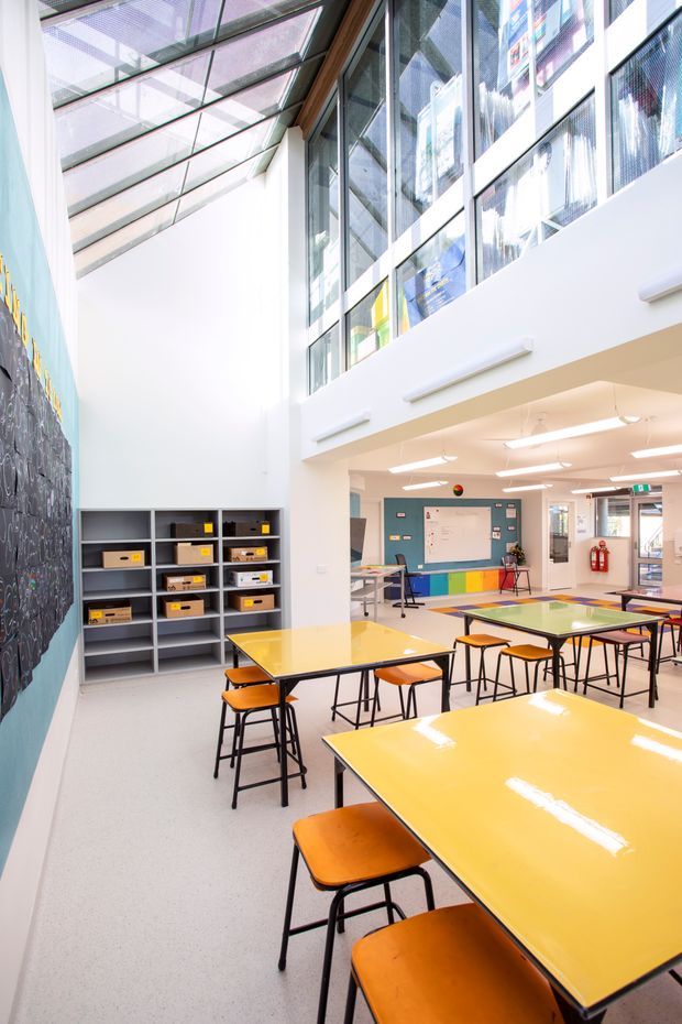 A bright contemporary school learning and dining area with numerous yellow rectangular tables and orange stools, open shelving with storage boxes, a large chalkboard-style wall, and a whiteboard at the rear. The double-height space has extensive upper-level glazing and a sloped glazed roof that floods the room with natural light. White walls, exposed dark window framing, colourful wall panels, and linear pendant lighting create a lively educational interior.