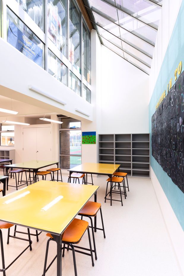 Bright contemporary school or educational learning space with a double-height atrium, clerestory glazing, and a glazed roof bringing in abundant natural light. The room contains multiple yellow rectangular tables with black metal legs and orange upholstered stools, open grey shelving, white walls, linear fluorescent lighting, and a large chalkboard wall with colourful graphic markings.