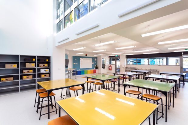 Bright, open-plan school classroom or learning commons with numerous rectangular tables and stools, open shelving with storage bins, a whiteboard, colourful wall panels, and extensive upper-level glazing. The space has a clean, functional institutional fit-out and a high ceiling with suspended linear fluorescent lighting.