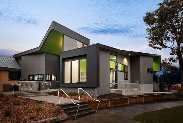 Modern multi-level building exterior at dusk, featuring dark grey cladding, bold angular roof forms, bright lime-green recessed facade accents, extensive glazing, and accessible entry stairs with metal handrails. A small paved terrace and landscaped foreground surround the structure.