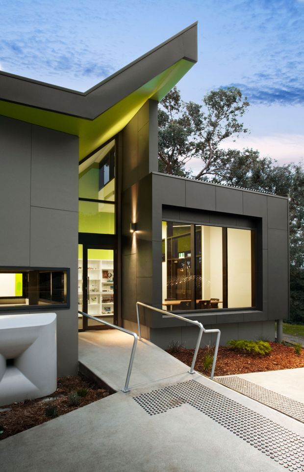 Exterior view of a contemporary building with crisp asymmetrical roof planes, dark grey panel cladding, large windows, and vivid lime-green soffit accents. A concrete accessible walkway with metal handrails leads to the glazed entrance, with a partially visible white exterior fixture in the foreground and landscaped planting around the building.