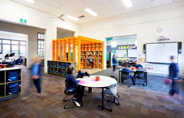 Bright contemporary primary-school classroom with students seated at round tables and workstations, an orange built-in shelving and reading/storage structure, whiteboards, educational displays, and large windows. The room has a patterned carpet floor, suspended acoustic ceiling, abundant fluorescent lighting, and an open connection to adjacent classroom areas.