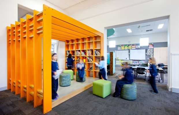 Bright contemporary school learning and library area with a striking orange timber-framed shelving and partition structure. The space includes integrated bookshelves, colourful upholstered ottomans, desks and computers, with students moving through the open-plan interior. White walls, suspended ceiling panels and grey carpet create a clean, practical educational setting.