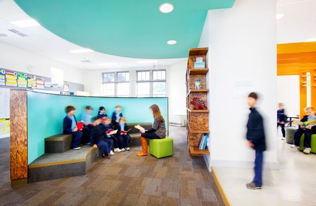 Bright contemporary school learning and reading area with children seated on tiered carpeted steps around a low turquoise partition. A teacher or adult sits on a vivid green cube seat, while open shelving displays books and objects. The space includes colorful wall graphics, large windows, white walls, a turquoise curved ceiling feature, and an adjacent orange-accented activity area.