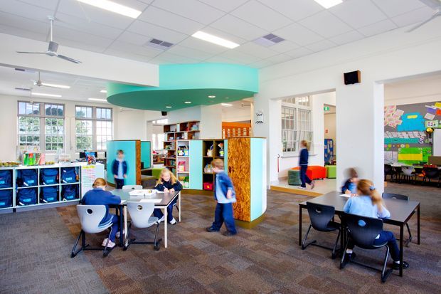 A bright, open-plan educational or childcare interior with children seated at tables, extensive shelving and storage, colourful artwork, and a distinctive suspended turquoise ceiling feature over a central collaborative area. The space has large multi-pane windows, acoustic ceiling tiles with fluorescent lighting, carpeted flooring, and glazed internal partitions connecting to adjacent activity rooms.