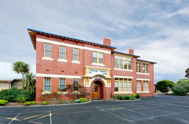 Exterior view of a historic two-storey red-brick school building with symmetrical classroom windows, cream-painted trim, arched central entrance, prominent school signage, chimneys, and a broad paved parking or play area in front. Established landscaping and mature trees surround the building.