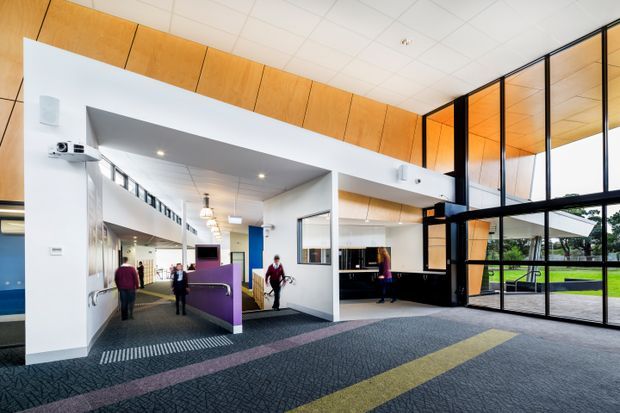 A bright, contemporary commercial interior lobby or reception area with an open circulation corridor, black-framed floor-to-ceiling glazing, white structural walls, warm timber wall and ceiling panelling, dark built-in reception joinery, and patterned carpet with coloured linear bands. Several people are present, and a landscaped outdoor area is visible through the glazing.