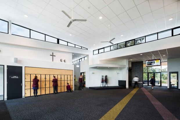 A bright, expansive institutional foyer or reception lobby with a high suspended grid ceiling, clerestory glazing, exposed ceiling fans, recessed lighting, and dark patterned carpet. A large bank of timber lockers lines the left wall beneath a cross, while a reception or circulation area with a dark tiled feature wall, glazed entry doors, and a yellow carpet pathway occupies the right side. People are present but not the focus.
