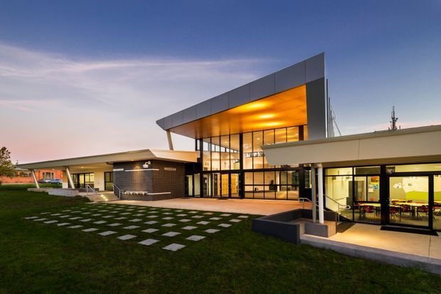 Exterior architectural photograph of a contemporary multi-wing building at dusk. The complex features a dramatic angular projecting roof canopy with a warm illuminated soffit, extensive floor-to-ceiling glazing, dark brick walls, and low flat-roofed wings. A paved entry terrace and stepping-stone path cross the landscaped lawn.