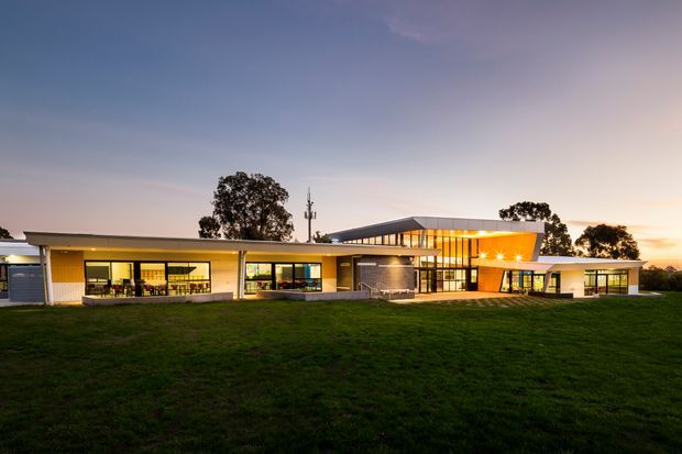 Wide dusk exterior view of a low-rise contemporary building set behind a broad lawn. The building has a long, horizontally composed form with flat and shallow-sloping roof planes, extensive glazed walls, warm interior lighting, dark framed windows, and a prominent elevated central volume with a glazed clerestory-like facade. Mature trees and a communications mast appear in the background.