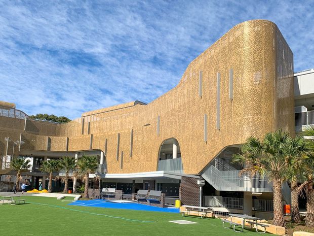 Exterior view of a large contemporary institutional or commercial building with a sculptural curved facade clad in a warm patterned material. Narrow vertical window openings, arched openings, exposed stair access, balconies, and extensive outdoor recreation or courtyard space with artificial turf and palm trees are visible under a bright blue sky.