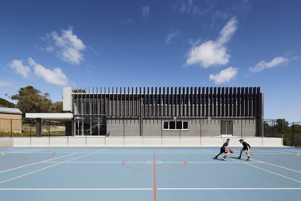 Exterior view of a contemporary multi-purpose sports or community building behind an outdoor basketball court. The building has a low, rectilinear form with a grey masonry lower level, extensive glazing, and a prominent upper facade of closely spaced vertical dark fins. The scene is brightly lit under a clear blue sky, with a blue marked court in the foreground.