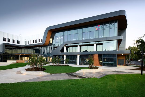 Exterior view of a contemporary multi-storey institutional or commercial building with a dramatic curved roofline and projecting upper-level forms. The facade combines extensive floor-to-ceiling glazing, dark metal panels, warm timber-toned feature cladding, and white structural elements. Landscaped lawns, paved paths, garden beds, and outdoor seating areas occupy the foreground.