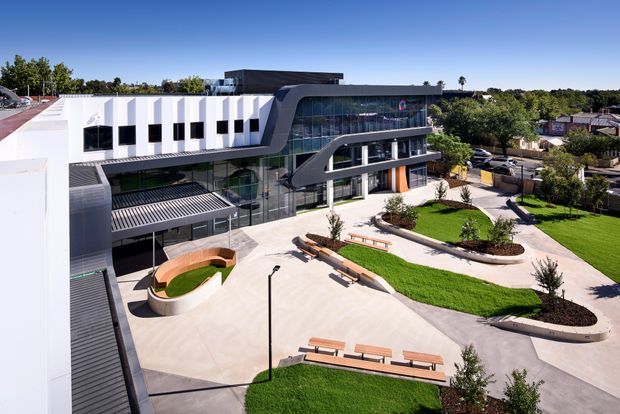 Aerial view of a contemporary multi-storey institutional or commercial building with a sculptural curved glazed facade, white rectilinear wings, dark metal roof and cladding elements, and landscaped forecourt. Paved paths, lawn areas, curved garden beds, benches, and an elliptical built-in seating feature surround the building.