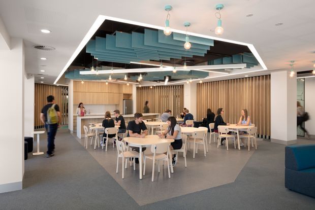 A bright contemporary commercial dining and breakout space with numerous round tables and white chairs, occupied by people, arranged beneath a striking suspended geometric ceiling feature. The ceiling incorporates layered teal acoustic panels, dark inset sections, linear lighting, and hanging pendant lights. Warm timber wall panelling and vertical timber slats surround the space, with carpeted flooring, white structural columns, and a partial blue upholstered seating area at right.