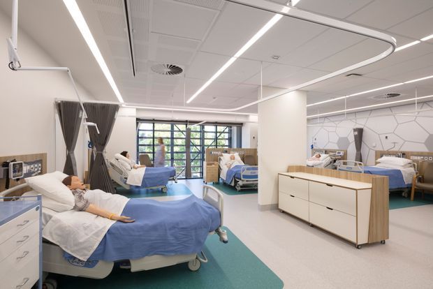 Bright contemporary hospital ward with multiple patient beds, white bedding, blue privacy curtains, pale timber storage cabinetry, turquoise flooring zones, and a white acoustic ceiling with linear integrated lighting. Large windows provide natural light, while a geometric hexagonal wall pattern adds a subtle feature to the clinical interior.