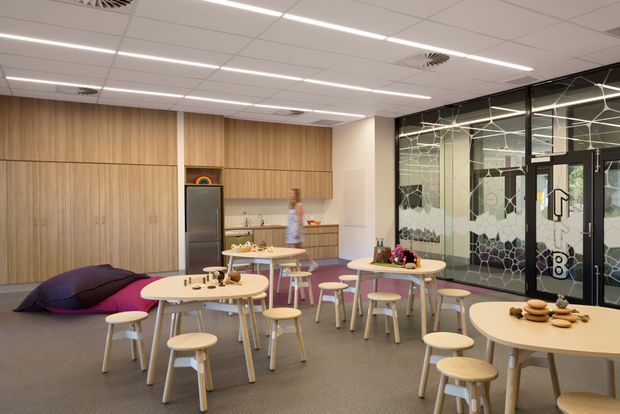 A bright contemporary communal breakout and dining area with multiple round tables, stools, and low beanbag seating. The space features extensive light timber wall cabinetry and panelled joinery, a compact kitchenette, grey carpet flooring, a suspended acoustic tile ceiling with linear lighting, and a glazed partition with decorative geometric patterned film. The arrangement and playful furnishings suggest a workplace, education, or commercial staff amenity area.