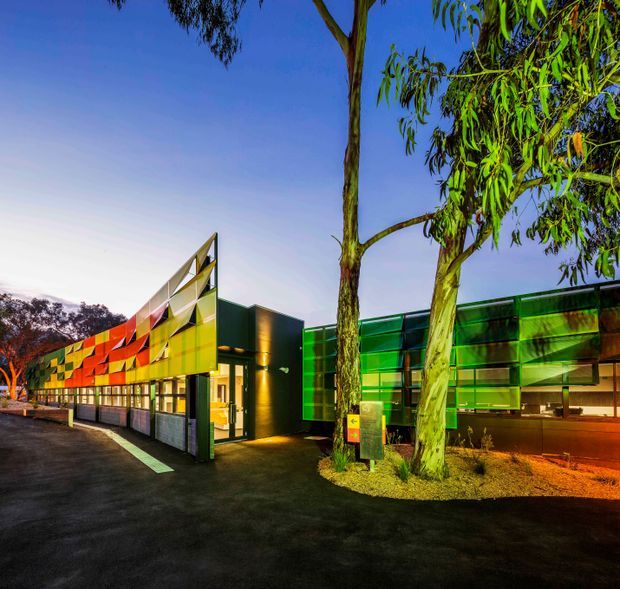 Night-time exterior view of a contemporary low-rise commercial or institutional building, featuring a strongly articulated angular entrance canopy and facade panels in vivid orange, yellow, green and translucent tones. Illuminated glazing and interior spaces are visible along the frontage, with mature eucalyptus trees and landscaped planting in the foreground.