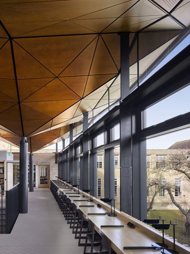 A long, contemporary institutional interior with a continuous timber study counter and rows of stools along a tall glazed wall. The space features exposed dark steel columns and framing, large windows, and a striking geometric perforated timber ceiling with triangular panel divisions. Brick and masonry walls are visible at the far end and through the glazing, creating a bright, refined study or workspace atmosphere.