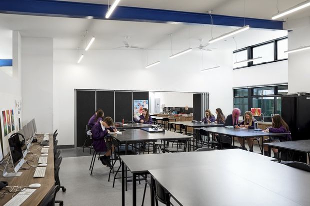 A bright, open-plan educational or commercial workspace with multiple large tables, desktop computers along a side bench, and people working or studying. The space has white walls, exposed blue structural beams, suspended linear lights, ceiling fans, large black-framed windows, and dark-framed folding or partition doors. The overall fit-out is functional, modern, and high-capacity.