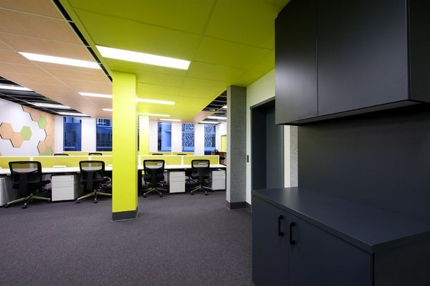 A bright contemporary open-plan office interior with rows of workstations, black office chairs, white desks, dark built-in cabinetry, grey carpet, and vivid lime-green ceiling and structural columns. Large windows and patterned acoustic wall panels add visual interest, while recessed linear lighting provides even illumination.