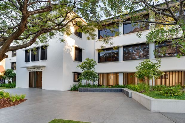 Exterior view of a contemporary multi-storey white rendered building with dark-framed windows, timber vertical screening, a recessed entry, landscaped planting, and a broad paved forecourt partially shaded by mature trees.