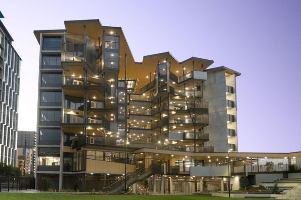 Exterior dusk view of a contemporary multi-storey apartment or mixed-use residential building, with articulated stepped massing, numerous projecting balconies, extensive glazing, dark metal railings, warm interior lighting, and a landscaped pedestrian entry at ground level. Adjacent urban buildings are visible at the edges.