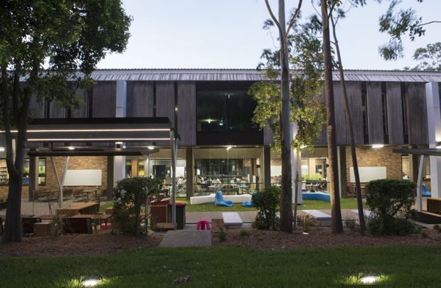 Exterior view of a contemporary two-storey building at dusk, with dark vertical timber cladding, extensive ground-floor glazing, covered outdoor seating areas, landscaped gardens, and illuminated interiors visible through the windows. Mature trees and low planting frame the facade.
