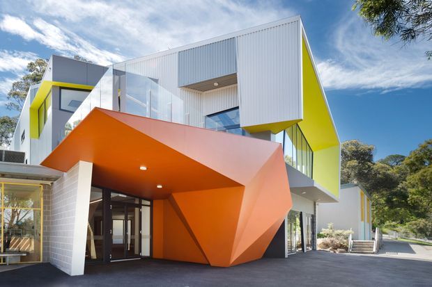 A striking contemporary multi-level building exterior with bold geometric forms. White and pale-grey corrugated cladding is contrasted by vivid lime-yellow wall reveals and a faceted orange angular canopy projecting over the glazed entrance. Large windows and transparent panels create a strong connection between interior and exterior, with paved forecourt and established trees surrounding the building.